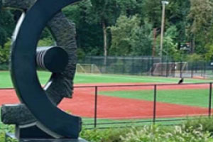 A dark, abstract circular sculpture stands in the foreground, with a baseball field visible behind a black metal fence. The field has an orange-red infield and a green turf outfield, backed by trees and a tall chain-link backstop.