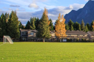 A wide, grassy sports field with a soccer goal on the left is backed by a row of townhouses and a mix of green and autumnal yellow trees. A large, dark mountain peak rises sharply on the right under a cloudy sky.